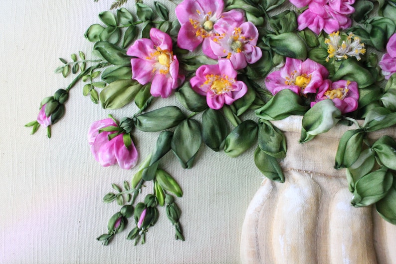 May include: A bouquet of pink silk ribbon roses in a cream-coloured vase. The roses are arranged in a cluster with green ribbon leaves. The background is a light green canvas.