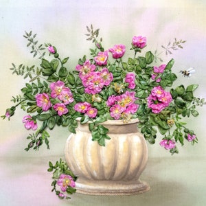 May include: A bouquet of pink silk ribbon flowers in a cream-coloured ceramic vase with green leaves. The vase has a round base and a fluted design.