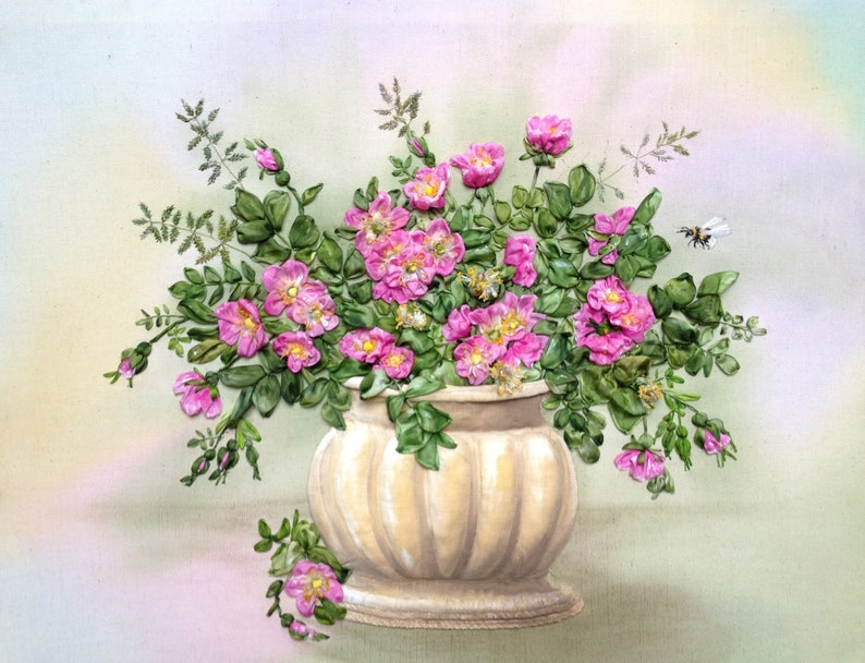 May include: A bouquet of pink silk ribbon flowers in a cream-coloured ceramic vase with green leaves. The vase has a round base and a fluted design.