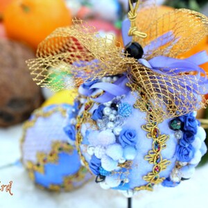 May include: A blue and gold decorative ornament with a net-like design and pearl-like accents. The ornament is decorated with blue and white flowers and is sitting on a white surface.