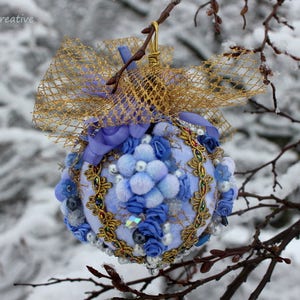 May include: A blue and gold Christmas bauble with a white bow and a gold mesh overlay. The bauble is decorated with blue and white flowers and beads. The bauble is hanging from a branch covered in snow.