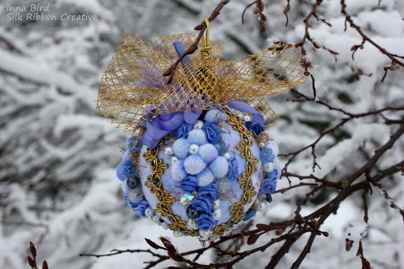 May include: A blue and gold Christmas bauble with a white bow and a gold mesh overlay. The bauble is decorated with blue and white flowers and beads. The bauble is hanging from a branch covered in snow.