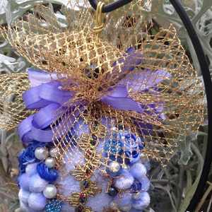 May include: A blue and gold Christmas bauble with a purple ribbon and a gold mesh bow. The bauble is decorated with blue and white beads, pearls, and fabric flowers.
