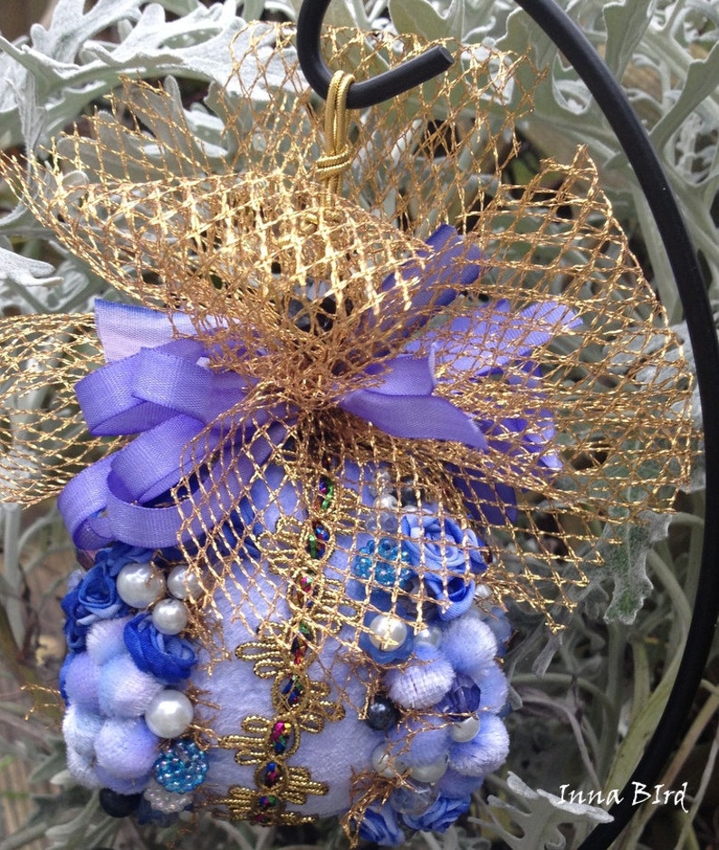 May include: A blue and gold Christmas bauble with a purple ribbon and a gold mesh bow. The bauble is decorated with blue and white beads, pearls, and fabric flowers.