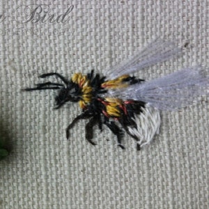 May include: A close-up of a hand-stitched bumblebee embroidered on a white fabric. The bumblebee has black and yellow stripes, white wings, and red accents.