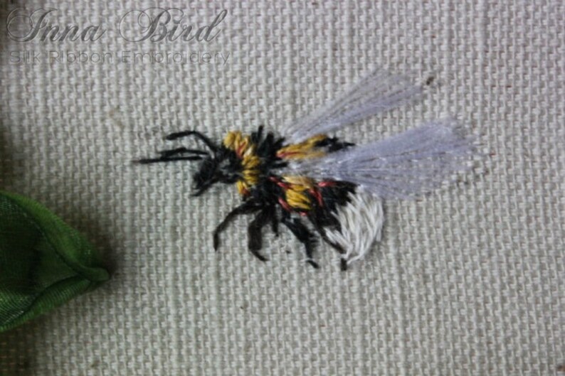 May include: A close-up of a hand-stitched bumblebee embroidered on a white fabric. The bumblebee has black and yellow stripes, white wings, and red accents.