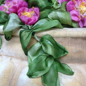 May include: A close-up of a silk ribbon embroidery of pink roses with green leaves. The embroidery is on a beige fabric background. The text "Silk Ribbon Embroidery" is visible in the top left corner.