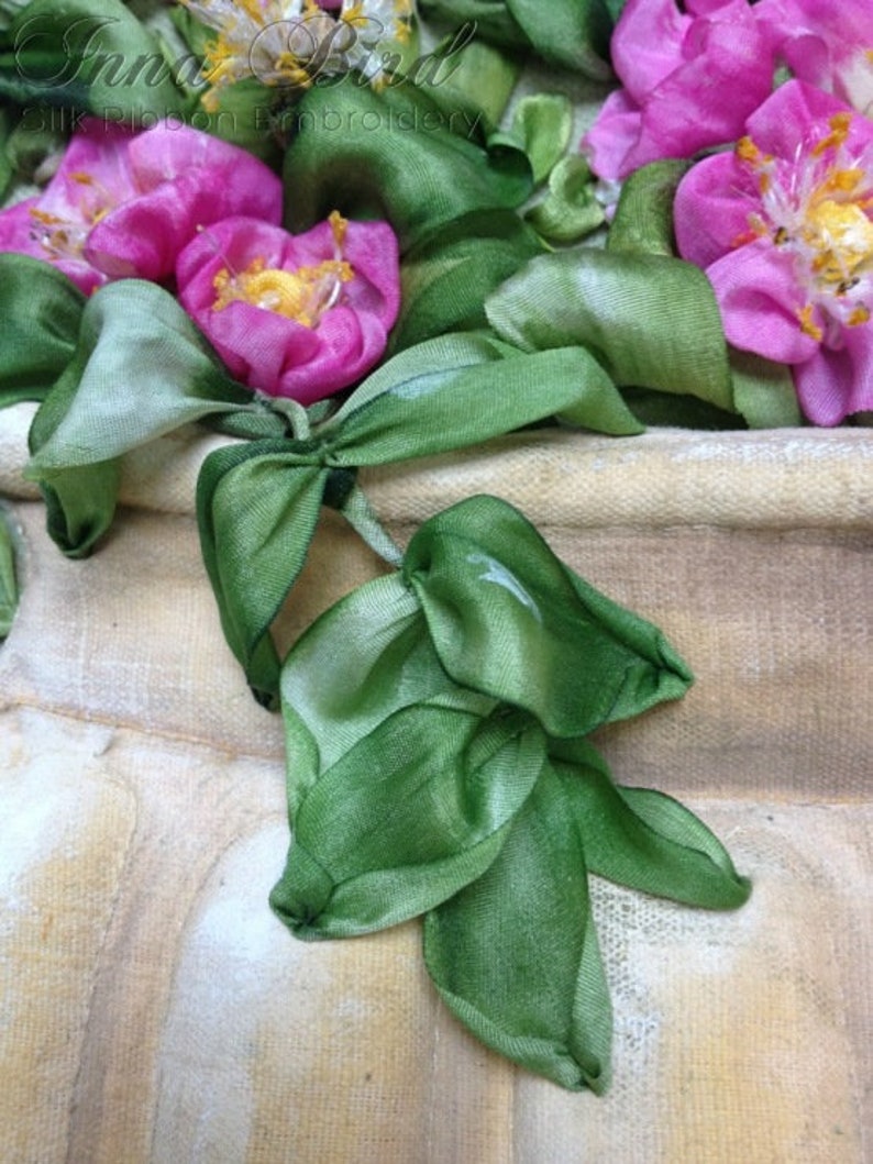 May include: A close-up of a silk ribbon embroidery of pink roses with green leaves. The embroidery is on a beige fabric background. The text "Silk Ribbon Embroidery" is visible in the top left corner.