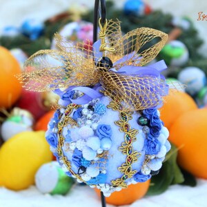 May include: A blue and gold Christmas bauble with a blue velvet base, decorated with white and blue rosettes, gold trim, and a gold mesh bow. The bauble is surrounded by oranges and other Christmas decorations.
