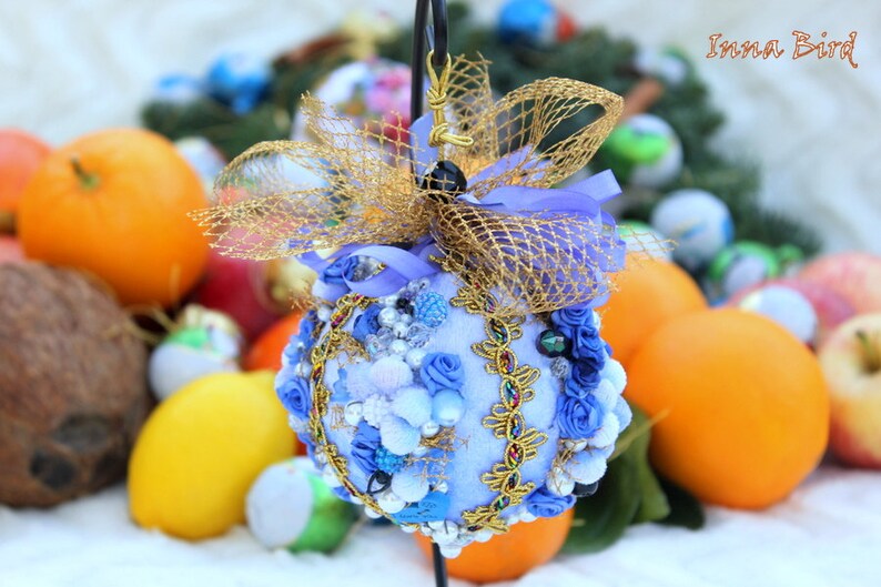 May include: A blue and gold Christmas bauble with a blue velvet base, decorated with white and blue rosettes, gold trim, and a gold mesh bow. The bauble is surrounded by oranges and other Christmas decorations.