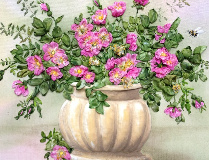 May include: A bouquet of pink silk ribbon roses in a cream-coloured ceramic vase with a ribbed design. The vase is sitting on a light green surface.