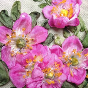 May include: Close-up of pink silk ribbon flowers with green leaves. The flowers are made with a technique called ribbon embroidery. The flowers have a delicate, feminine look.