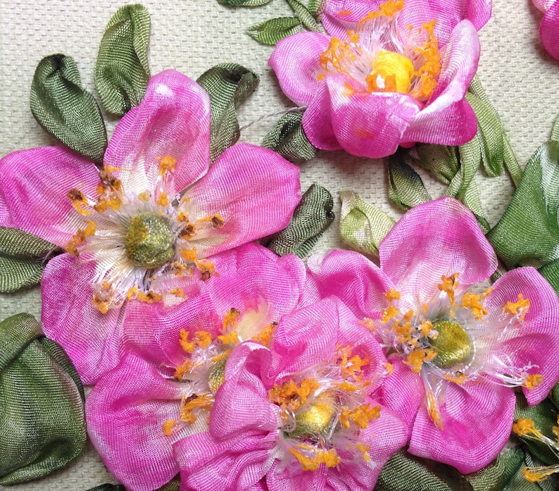 May include: Close-up of pink silk ribbon flowers with green leaves. The flowers are made with a technique called ribbon embroidery. The flowers have a delicate, feminine look.