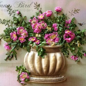 May include: A bouquet of pink silk ribbon flowers in a beige ceramic vase with a ribbed design. The flowers are arranged in a vase with a base and are surrounded by green silk ribbon leaves. The background is a light pink and green gradient.