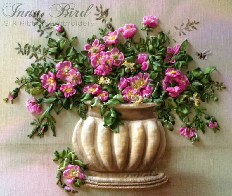 May include: A bouquet of pink silk ribbon flowers in a beige ceramic vase with a ribbed design. The flowers are arranged in a vase with a base and are surrounded by green silk ribbon leaves. The background is a light pink and green gradient.