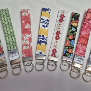 May include: A selection of fabric key fobs with diverse patterns, including polka dots, floral, and geometric designs. Each fob has a metal clasp and a tag reading "Duchess Handmade By". The key fobs are in various colours, such as blue, pink, and green.