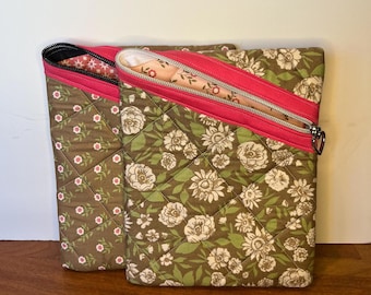 May include: Two quilted fabric zippered pouches with floral patterns. The pouch on the left has a green background with white flowers and pink polka dots. The pouch on the right has a green background with white flowers and green leaves. Both pouches have pink zippers.