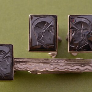 May include: A set of silver cufflinks and a tie bar featuring black onyx stones with engraved images of a person wearing a helmet.