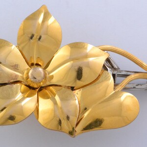 May include: A gold-toned flower brooch with a silver-toned clasp. The brooch is made of metal and has a floral design. The flower has five petals and a center.
