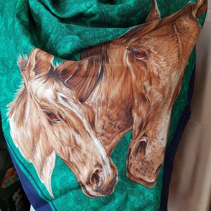 Puede incluir: Un pañuelo cuadrado con una ilustración detallada de dos caballos marrones con ojos rojos. El pañuelo tiene un estampado de cachemira verde y un borde azul oscuro. El pañuelo está hecho de una tela suave y fluida.