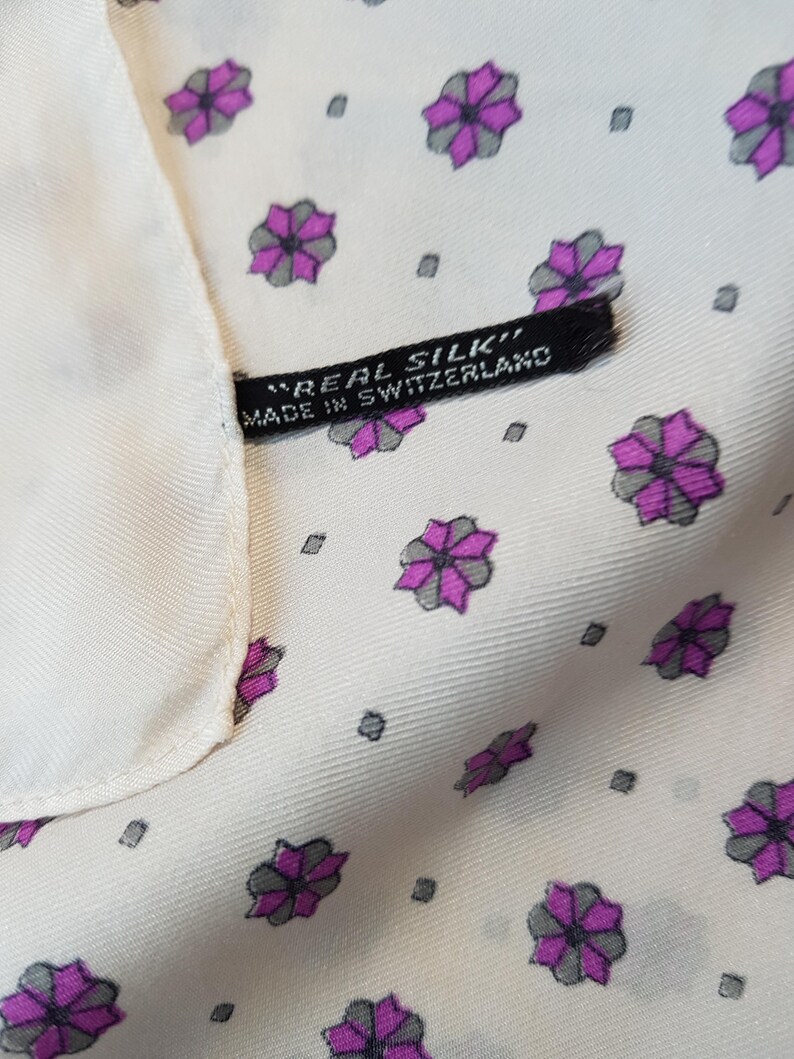 Może przedstawiać: Biały jedwabny szal z powtarzającym się wzorem małych fioletowych i szarych kwiat&oacute;w. Szal ma czarną metkę z napisem "Real Silk" i "Made in Switzerland".