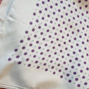 Może przedstawiać: Biała bawełniana bandana z fioletowym wzorem kwiatowym. Bandana jest złożona w kształt tr&oacute;jkąta.
