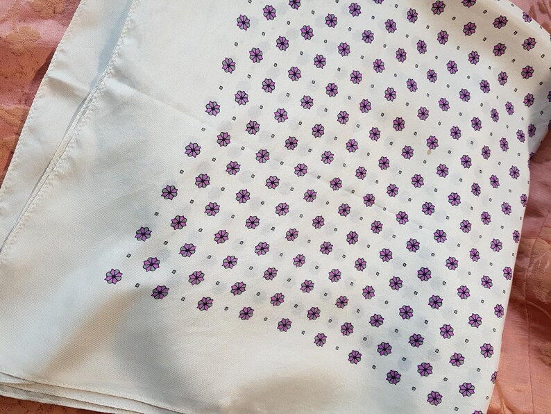Może przedstawiać: Biała bawełniana bandana z fioletowym wzorem kwiatowym. Bandana jest złożona w kształt tr&oacute;jkąta.