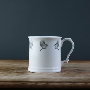 Puede incluir: Una taza de cerámica blanca con un diseño de pájaro negro. La taza tiene un asa y una base redondeada.