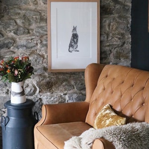 Puede incluir: Un dibujo en blanco y negro enmarcado de un conejo sentado, colgado en una pared de piedra sobre un sillón de cuero marrón con una manta y una almohada dorada.
