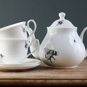 May include: A white teapot and two teacups with matching saucers. The teapot and cups are decorated with black and white bee illustrations.