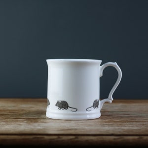 Puede incluir: Taza de cerámica blanca con asa decorativa. La taza presenta un patrón repetido de pequeños ratones grises alrededor de la base. La taza está colocada sobre un fondo oscuro sobre una superficie de madera.