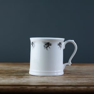 May include: White ceramic mug with a black and white bee design. The mug has a handle and is sitting on a wooden surface.