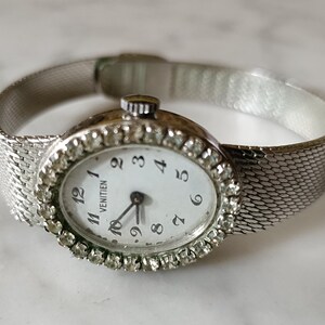 Pode incluir: Um relógio prateado com um mostrador branco e uma pulseira de malha prateada. O mostrador é rodeado por strass e tem marcadores de hora em algarismos romanos pretos. O mostrador do relógio também tem a palavra "VENTIEN" impressa.