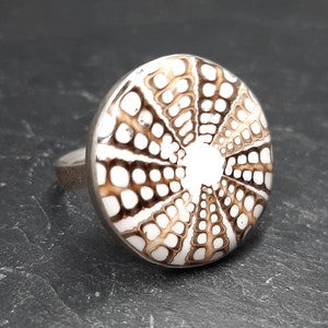 Könnte beinhalten: Ein silberner Ring mit einem weißen und braunen Muster. Das Muster ähnelt einem Seeigel.