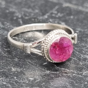 May include: A silver ring with a pink gemstone set in a bezel setting. The ring has a braided band and a split shank.