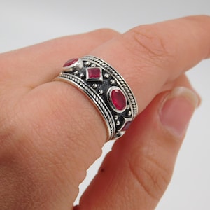 Peut inclure: Bague en argent ornée de pierres précieuses rouges. La bague présente un motif de bande avec des pierres rouges ovales et carrées alternées, entourées d'une bordure texturée détaillée. La bague est portée au doigt.