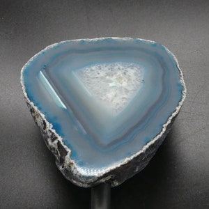 Pode incluir: Uma fatia de geoda de ágata azul com um centro branco e uma borda externa cinza escura. A geoda está montada em um suporte preto.