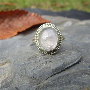 Puede incluir: Un anillo de plata con una piedra preciosa ovalada transparente engastada en un engaste de cuentas. El anillo está sobre una superficie de roca gris.