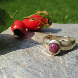 Könnte beinhalten: Ein silberner Ring mit einem rosafarbenen Edelstein, der in einer Zargenfassung gefasst ist. Der Ring liegt auf einem grauen Stein mit roten Beeren im Hintergrund.