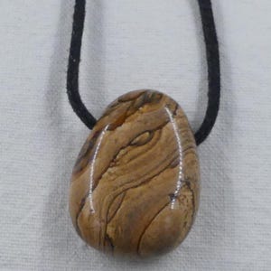 Pode incluir: Um pingente de pedra marrom e bege com um padr&atilde;o de redemoinho, pendurado em um cord&atilde;o preto.