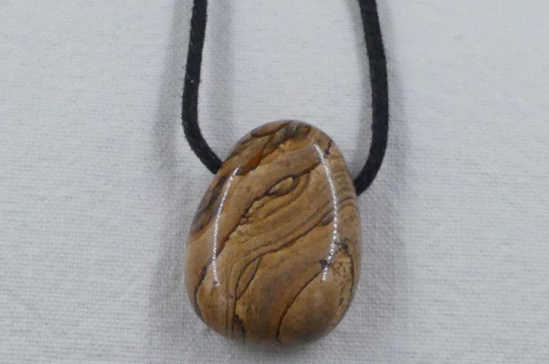 Pode incluir: Um pingente de pedra marrom e bege com um padr&atilde;o de redemoinho, pendurado em um cord&atilde;o preto.