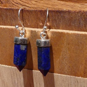 Könnte beinhalten: Ein Paar silberne Ohrringe mit blauen Lapislazuli-Tropfen. Die Ohrringe hängen an einer Holzoberfläche.