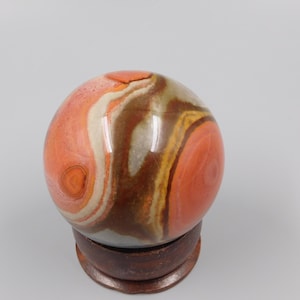Op de afbeelding: Een gepolijste, bolvormige steen met wervelende banden in terracotta oranje, donkerbruin, crème en gedempt grijs. De gladde, ronde minerale bol rust stevig in een cirkelvormige, donkerbruine houten displaystandaard met een gepolijste afwerking.