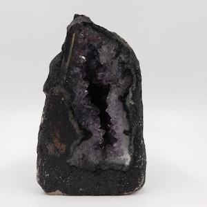 Könnte beinhalten: Eine dunkle, rau strukturierte Geode mit einem Hohlraum, der mit funkelnden lila Kristallen ausgekleidet ist. Die Außenseite ist tiefschwarz und kontrastiert mit dem helleren Inneren. Dieses Naturstein-Exemplar ist ein einzigartiger Dekorationsartikel.