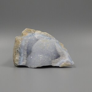Könnte beinhalten: Eine natürliche Geode aus blauem Spitzenachat. Die Geode hat ein hellblaues kristallines Inneres und eine raue, beige Außenseite. Der Stein wird auf grauem Hintergrund präsentiert und zeigt seine natürliche Form und Textur. Ein einzigartiges Exemplar für Sammler.