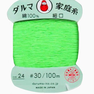 May include: A spool of green cotton thread labeled "Daruma Family Thread" with the text "Cotton 100%" and "Fine Mouth". The spool is labeled with the number "24" and "#30/100m".