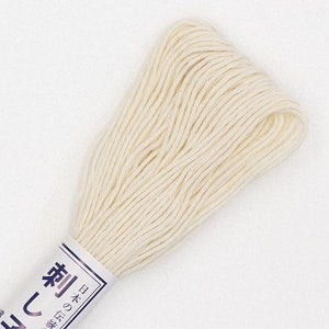 May include: A skein of off-white embroidery floss. The floss is tightly wound and secured with a paper label featuring Japanese text. The thread appears soft and ready for use in various needlework projects.