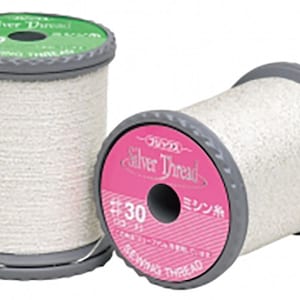 May include: Two spools of silver sewing thread. The thread is wound on gray plastic spools with green and pink labels. The labels read "Silver Thread" and include Japanese text. The thread is a light, shimmering color.