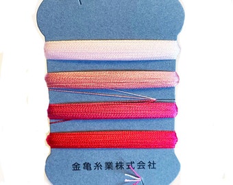 May include: A blue card holds six spools of thread in shades of pink and red. The card has a curved top with a slit and Japanese text at the bottom. The threads are neatly wound, with a few strands pulled out.