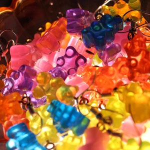 Pu&ograve; includere: Orecchini a forma di orsetti gommosi colorati in varie tonalit&agrave; di arancione, giallo, rosa, viola e blu. Gli orecchini sono in plastica traslucida e hanno ganci color argento. Gli orecchini sono esposti in una ciotola di vetro decorativa.